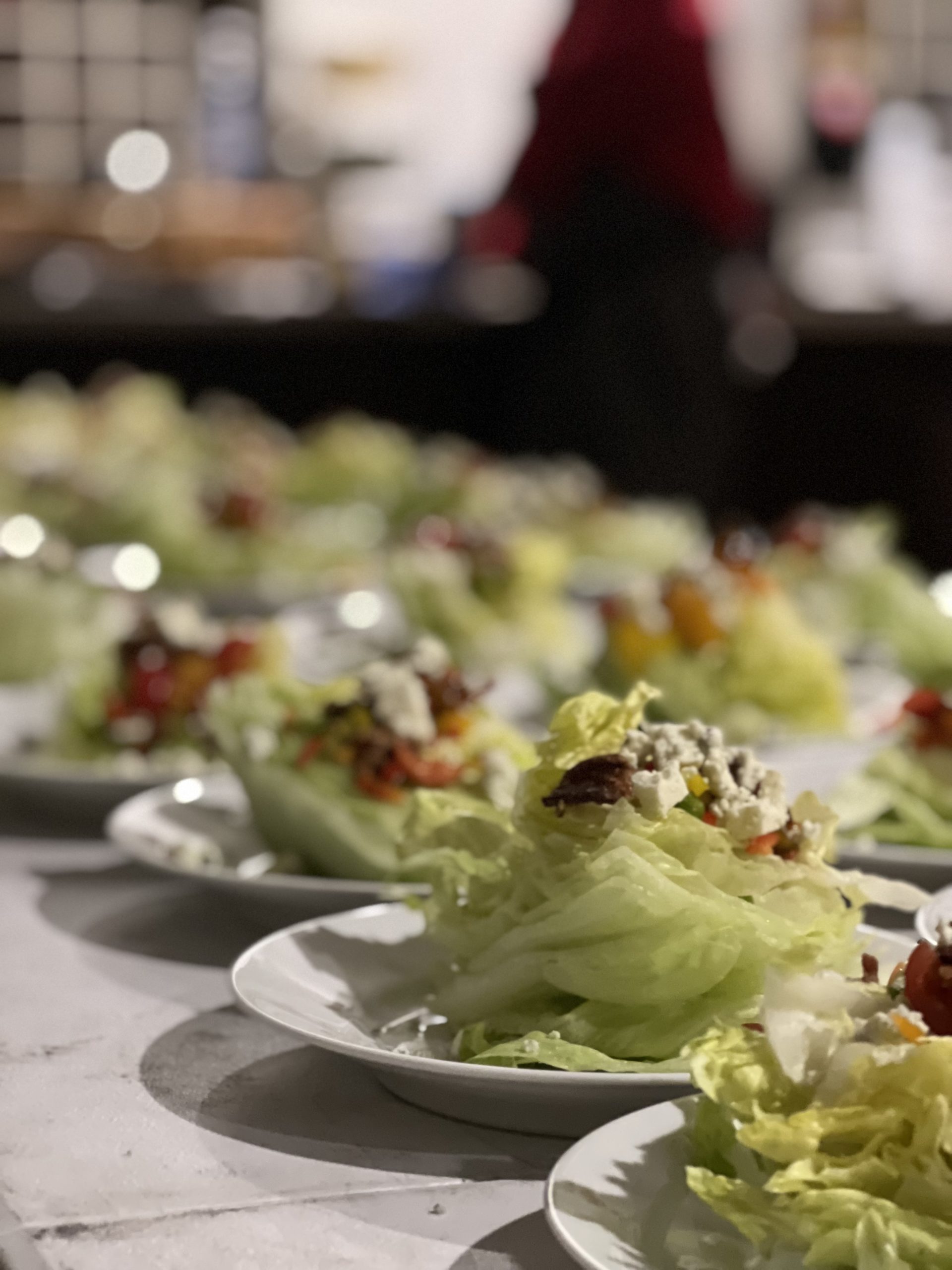 A beautifully arranged row of plated wedge salads featuring crisp iceberg lettuce, crumbled blue cheese, cherry tomatoes, bacon bits, and a drizzle of dressing, set on a marble countertop. A blurred background shows a catering setup with a server in a red uniform.