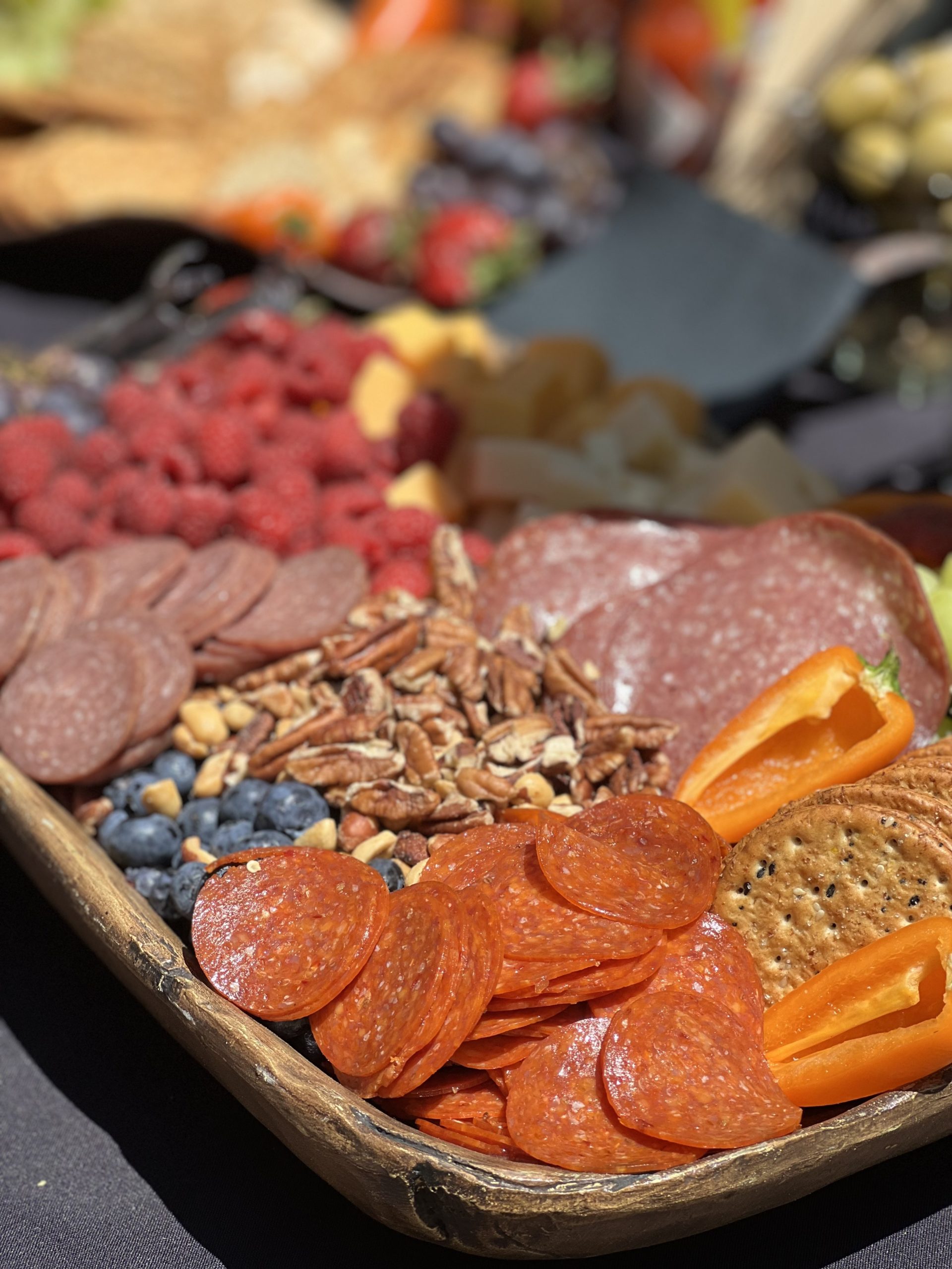 A beautifully arranged charcuterie board featuring an assortment of cured meats, fresh raspberries, blueberries, mixed nuts, sliced bell peppers, and crackers. The board is set on a rustic wooden platter with a blurred background of additional appetizers and garnishes.